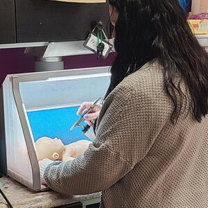 Puede incluir: Una persona con un c&aacute;rdigan beige texturizado utiliza un aer&oacute;grafo plateado para pintar una peque&ntilde;a cabeza de mu&ntilde;eca de piel clara dentro de una cabina de pintura port&aacute;til e iluminada con un panel trasero azul brillante.