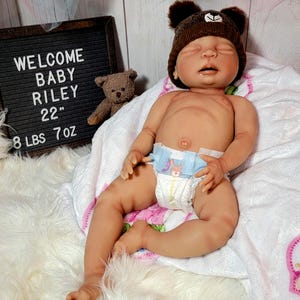 May include: A realistic newborn doll with light skin, wearing a brown bear-ear hat and a disposable diaper. A small brown teddy bear sits next to a black letter board that reads: WELCOME BABY RILEY 22" 8 LBS 7 OZ. The doll rests on a white blanket.