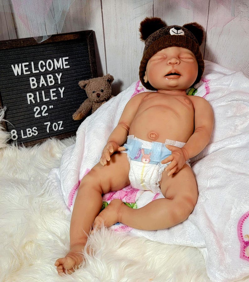 May include: A realistic newborn doll with light skin, wearing a brown bear-ear hat and a disposable diaper. A small brown teddy bear sits next to a black letter board that reads: WELCOME BABY RILEY 22" 8 LBS 7 OZ. The doll rests on a white blanket.