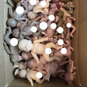 May include: A cardboard box filled with numerous small, unpainted baby doll parts in various skin tones. The dolls are in different poses, some with arms outstretched, others curled up.