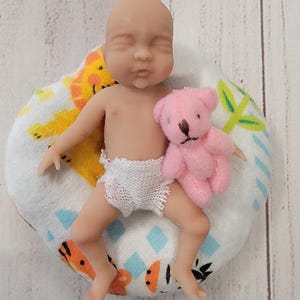 Puede incluir: Una figura de beb&eacute; en miniatura con los ojos cerrados, que lleva un pa&ntilde;al de ganchillo blanco, acostada sobre un coj&iacute;n de tela blanca con ilustraciones de animales en naranja y amarillo, sosteniendo un peque&ntilde;o osito de peluche rosa.