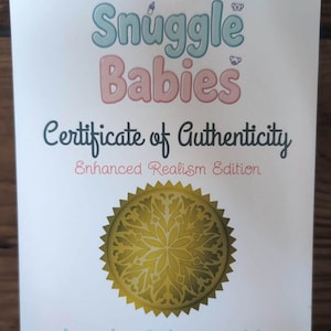May include: A white certificate of authenticity for Silicone Snuggle Babies, Enhanced Realism Edition. It features pink and blue text, a gold circular seal with a floral pattern, and the name Jennifer Schoenwalder, Silicone Doll Artist.