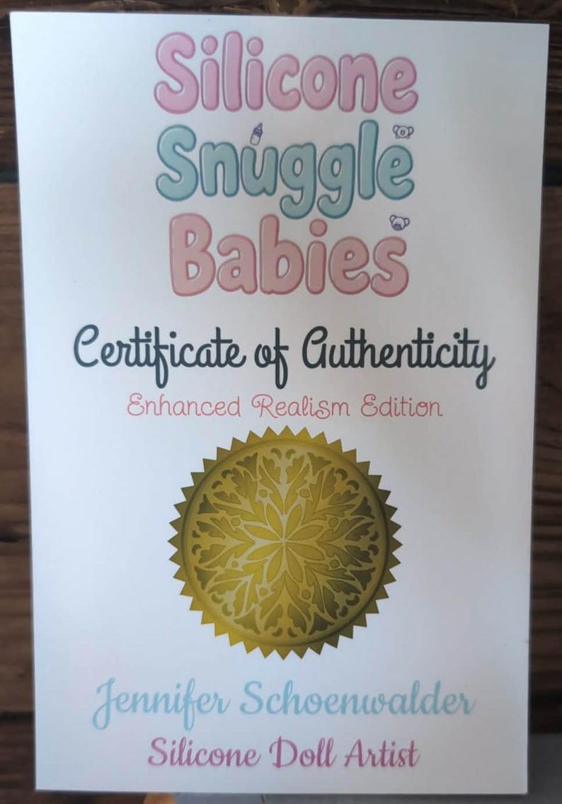 May include: A white certificate of authenticity for Silicone Snuggle Babies, Enhanced Realism Edition. It features pink and blue text, a gold circular seal with a floral pattern, and the name Jennifer Schoenwalder, Silicone Doll Artist.