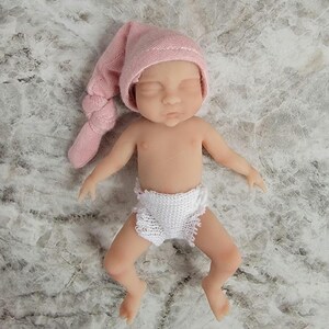 Puede incluir: Una peque&ntilde;a mu&ntilde;eca de silicona realista con piel clara, ojos cerrados y expresi&oacute;n neutra. La mu&ntilde;eca lleva un gorro de punto suave de color rosa p&aacute;lido con un extremo anudado y un pa&ntilde;al de punto blanco texturizado. La figura est&aacute; acostada.
