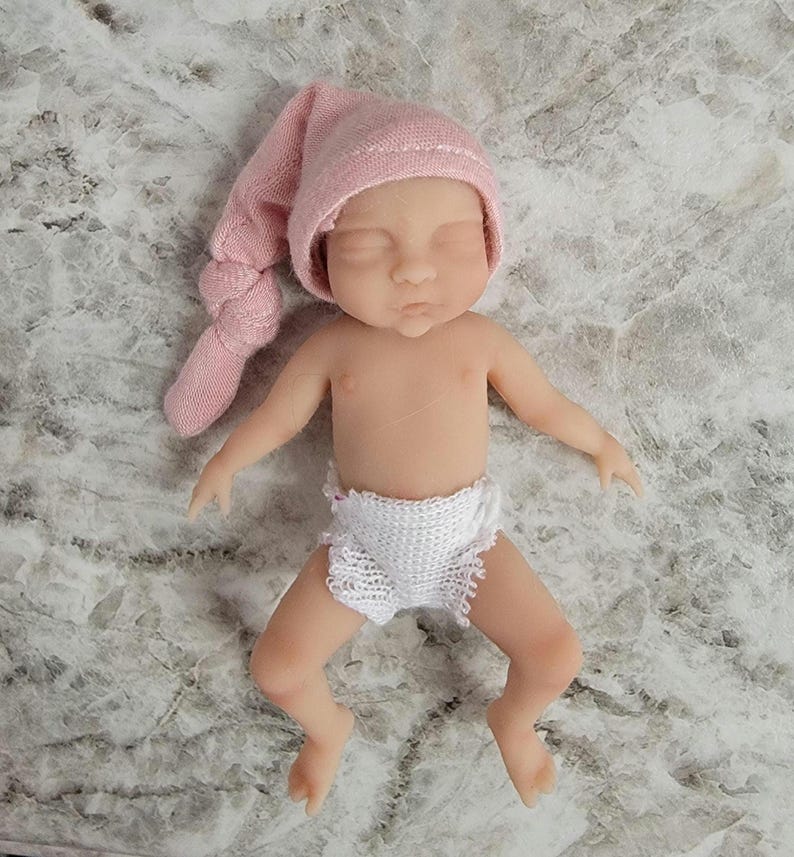 Puede incluir: Una peque&ntilde;a mu&ntilde;eca de silicona realista con piel clara, ojos cerrados y expresi&oacute;n neutra. La mu&ntilde;eca lleva un gorro de punto suave de color rosa p&aacute;lido con un extremo anudado y un pa&ntilde;al de punto blanco texturizado. La figura est&aacute; acostada.