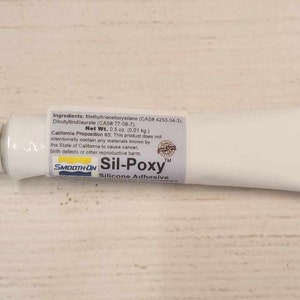 Może przedstawiać: Biała tubka kleju silikonowego Sil-Poxy z czarną i żółtą etykietą. Etykieta zawiera napis "Sil-Poxy Silicone Adhesive" i "Smooth-On". Tubka ma pojemność 0,5 uncji (0,01 kg).
