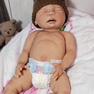 May include: A realistic newborn baby doll with closed eyes, wearing a brown knit hat with two small pom-poms resembling bear ears. The doll is wearing a disposable diaper with a small graphic print on the front and is resting on a white blanket.