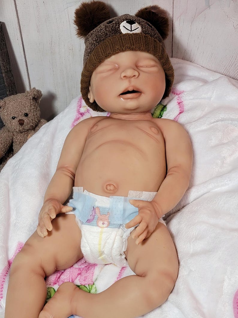 May include: A realistic newborn baby doll with closed eyes, wearing a brown knit hat with two small pom-poms resembling bear ears. The doll is wearing a disposable diaper with a small graphic print on the front and is resting on a white blanket.