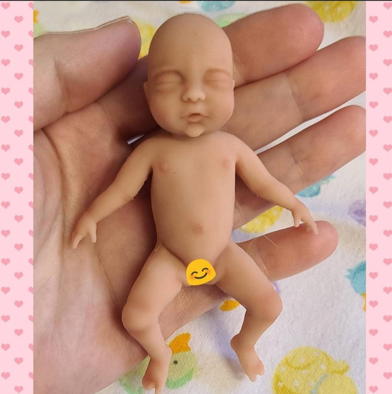 Puede incluir: Una mu&ntilde;eca beb&eacute; en miniatura de piel clara con un acabado suave y mate. La mu&ntilde;eca presenta una cabeza calva, ojos cerrados, una nariz peque&ntilde;a y una boca ligeramente abierta. Tiene dedos y pies definidos, y una peque&ntilde;a pegatina de emoji amarilla cubre el &aacute;rea p&eacute;lvica.