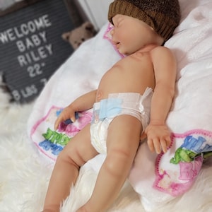 May include: A realistic reborn baby doll with light skin, wearing a brown knit beanie with a pom-pom and a disposable diaper. The doll is lying on a white blanket with colorful graphics, positioned near a letter board that reads WELCOME BABY RILEY.