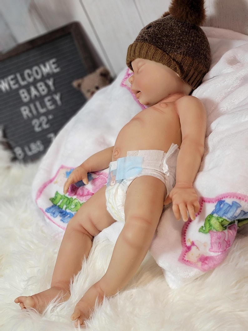 May include: A realistic reborn baby doll with light skin, wearing a brown knit beanie with a pom-pom and a disposable diaper. The doll is lying on a white blanket with colorful graphics, positioned near a letter board that reads WELCOME BABY RILEY.