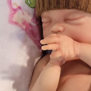 May include: A realistic newborn doll with closed eyes and pale skin, wearing a brown knit beanie with a white bear face patch. The doll is positioned with one hand near its mouth, resting against a white fabric surface featuring a pink and green graphic.