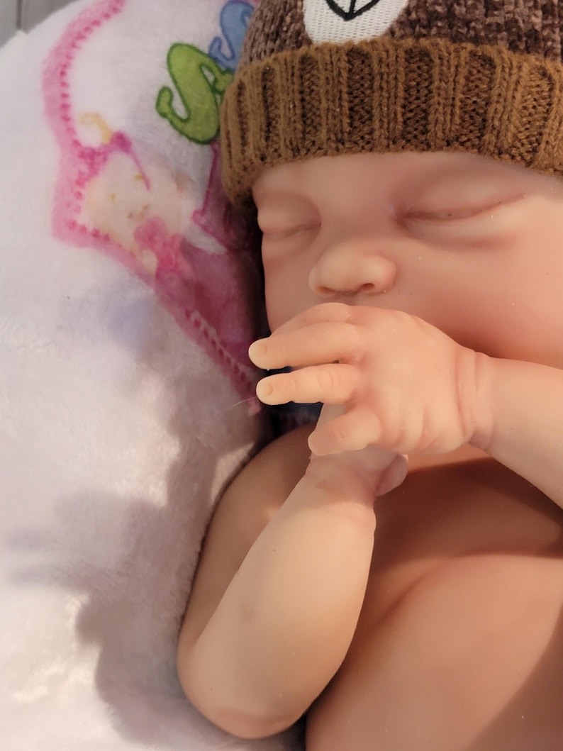 May include: A realistic newborn doll with closed eyes and pale skin, wearing a brown knit beanie with a white bear face patch. The doll is positioned with one hand near its mouth, resting against a white fabric surface featuring a pink and green graphic.