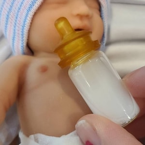 以下が含まれることがあります： ピンクとブルーのストライプの帽子をかぶったシリコン製の赤ちゃん人形で、口に小さなミルク瓶を持っています。