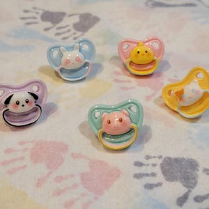 May include: A collection of baby pacifiers in various pastel colors. Each pacifier features a different animal design, including a bunny, chick, dog, pig, and cat. The pacifiers are made of plastic and have a heart-shaped shield.