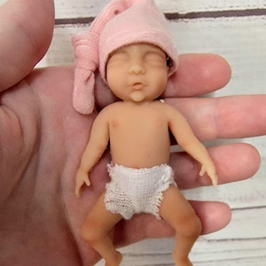 Puede incluir: Una mu&ntilde;eca en miniatura de piel clara, con los ojos cerrados y una boca peque&ntilde;a. La mu&ntilde;eca lleva un gorro de punto rosa claro suave con un detalle anudado en el lateral y un pa&ntilde;al de ganchillo blanco con textura. La figura tiene un acabado mate liso.