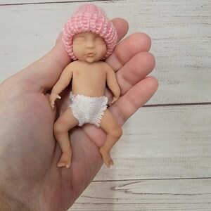 Puede incluir: Una figura de beb&eacute; en miniatura con una tez clara y suave, que lleva un gorro de punto rosa suave y un pa&ntilde;al de ganchillo blanco. La peque&ntilde;a mu&ntilde;eca tiene los ojos cerrados, una expresi&oacute;n neutra y se muestra en posici&oacute;n de descanso.
