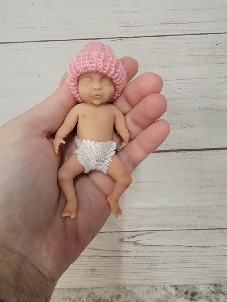 Puede incluir: Una figura de beb&eacute; en miniatura con una tez clara y suave, que lleva un gorro de punto rosa suave y un pa&ntilde;al de ganchillo blanco. La peque&ntilde;a mu&ntilde;eca tiene los ojos cerrados, una expresi&oacute;n neutra y se muestra en posici&oacute;n de descanso.