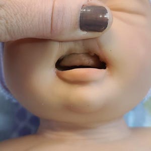 May include: A realistic baby doll with smooth, light-toned skin, closed eyes, and a slightly open mouth revealing a tongue. The doll wears a soft, light-colored knit cap with subtle blue and white stripes, covering the top of its head.