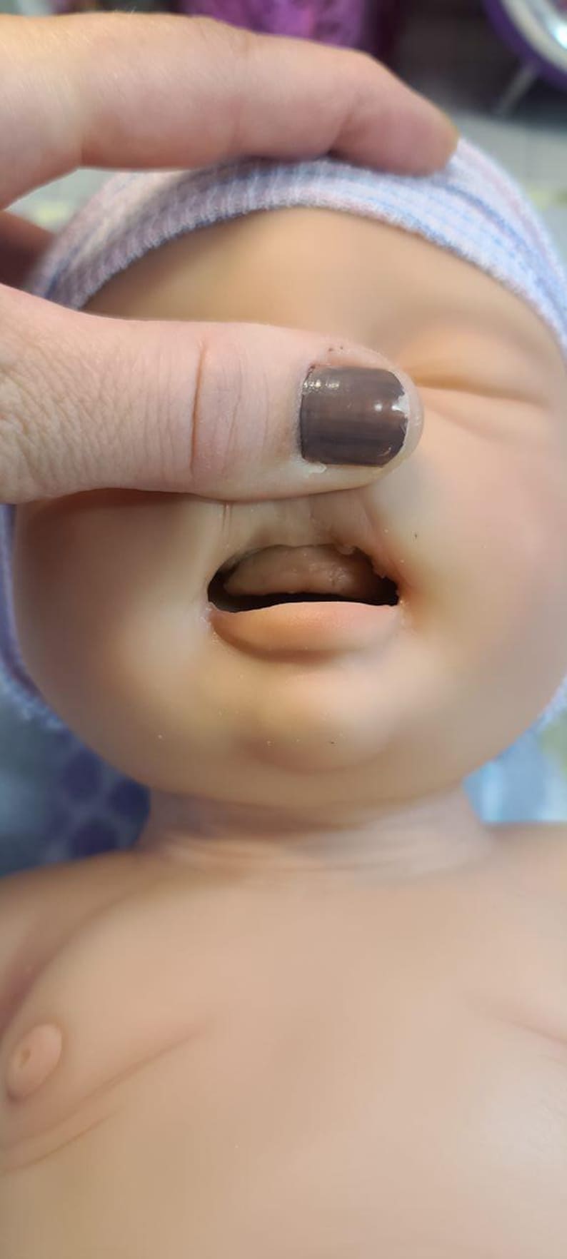 May include: A realistic baby doll with smooth, light-toned skin, closed eyes, and a slightly open mouth revealing a tongue. The doll wears a soft, light-colored knit cap with subtle blue and white stripes, covering the top of its head.