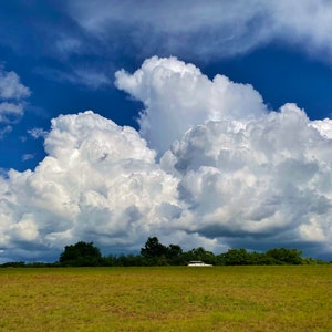 Può includere: Una grande nube bianca cumuliforme riempie il cielo sopra un campo verde. La nube è soffice e ondulata, con una parte inferiore grigio scuro. Il campo è ricoperto di erba verde corta.