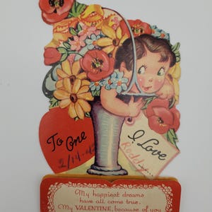 May include: A vintage Valentine's Day card featuring a girl peeking out from behind a vase of flowers. The card has a red heart with the text "To One" and the date "2/14-44". The card also has a tag that says "I Love" with a name written below. The card has a message that reads "My happiest dreams have all come true. My VALENTINE, because of you."