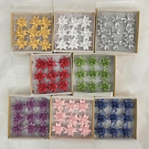 May include: A collection of handmade paper star ornaments in various colors, including gold, white, silver, red, green, purple, pink, and blue. Each set of stars is neatly arranged in a small, square cardboard box, perfect for crafting or holiday decorations.