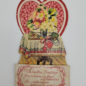 May include: Vintage Valentine's Day card featuring a heart-shaped frame with a floral arrangement, a table setting with a vase, a fan, a ribbon, and a necklace. The card reads "Valentine Greetings Your charming Grace, my Valentine Has captured me, Will you be mine?"