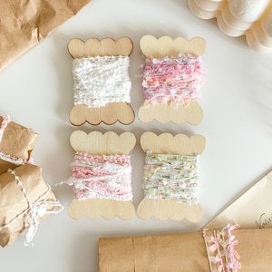 Peut inclure: Quatre bobines de fil coloré dans des supports en bois. Le fil est blanc, rose, rose clair et vert. Les bobines sont disposées en deux rangées de deux.