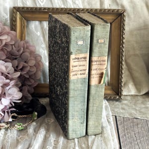May include: Two vintage books with faded blue covers and labels, standing upright. The books are titled "Biographie Universelle" and have aged, textured spines. A gold-framed picture and pink flowers are in the background.