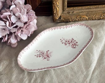 May include: White oval serving dish with a maroon floral and bird design. The dish has a maroon border and is set on a neutral-coloured surface. The dish is surrounded by a pink flower arrangement and antique picture frames.