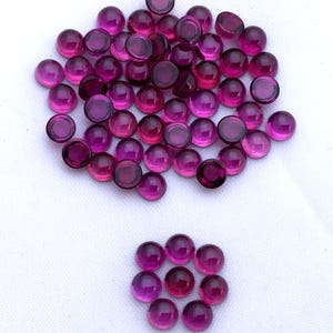 Puede incluir: Una gran pila de piedras preciosas redondas y moradas en cabujón, y un grupo más pequeño de las mismas piedras preciosas dispuestas en círculo.