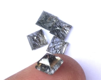 May include: Four clear quartz stones with black inclusions, also known as rutilated quartz, in a square shape. The stones are on a finger and a white surface.