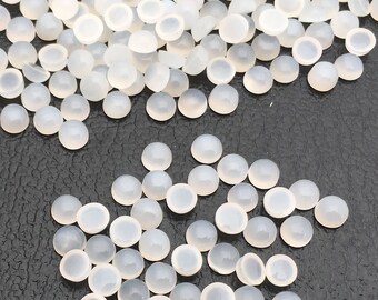 May include: A collection of small, round, translucent cabochon gemstones. The gemstones are a milky white color and have a smooth, polished surface. They are scattered across a dark surface, creating a contrast.
