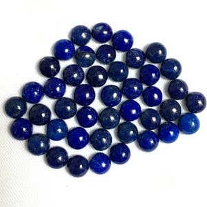 May include: A collection of round, dark blue lapis lazuli gemstone beads. The beads are smooth and polished, and they are arranged in a loose pile.