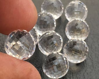 May include: Eight clear faceted crystal balls, some with a slight rainbow effect, are arranged on a dark gray surface. One hand is holding one of the balls.