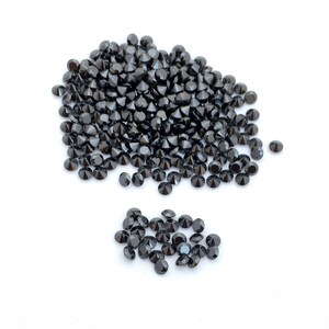 May include: A pile of black round gemstones, likely diamonds, on a white surface. The gemstones are arranged in a loose pile, with some spilling out to the side.