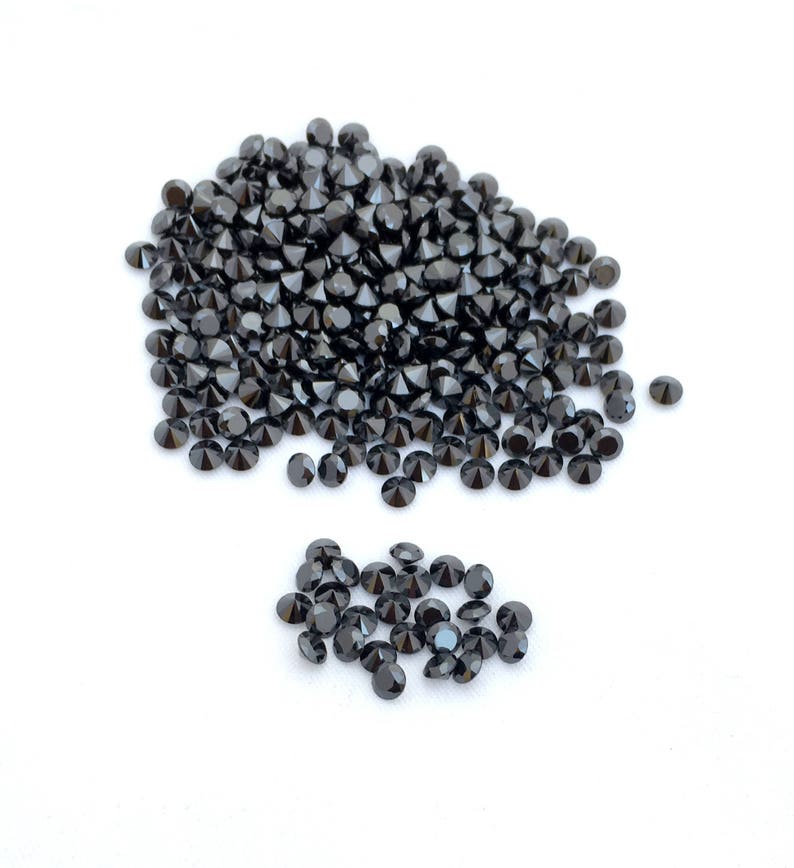 May include: A pile of black round gemstones, likely diamonds, on a white surface. The gemstones are arranged in a loose pile, with some spilling out to the side.
