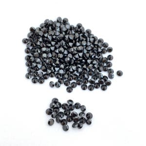 May include: A large pile of black round gemstones, with a smaller pile of the same gemstones to the side. The gemstones are scattered on a white surface.
