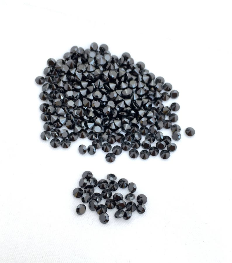 May include: A large pile of black round gemstones, with a smaller pile of the same gemstones to the side. The gemstones are scattered on a white surface.