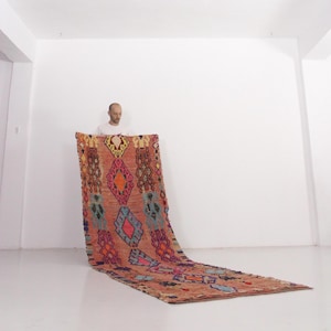 May include: A long, rectangular rug featuring a terracotta-colored base with a repeating pattern of geometric diamond shapes in vibrant shades of blue, pink, orange, and yellow. The rug is displayed vertically, extending from a raised position to the floor.