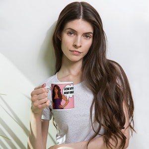 May include: White ceramic mug with a pink background and a cartoon image of a woman with long brown hair wearing a purple dress. The woman is holding a speech bubble that says "Gone with the wind, fabulously".
