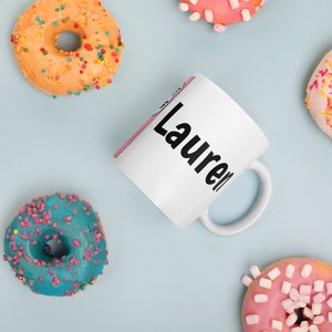 May include: White ceramic mug with the name "Lauren" written in black lettering. The mug is surrounded by colorful donuts with sprinkles.
