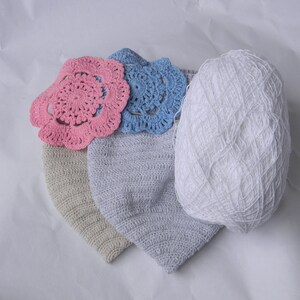 May include: Two crocheted hats, one beige and one light blue, with a pink and a blue crocheted flower on top. A ball of white yarn is also in the image.