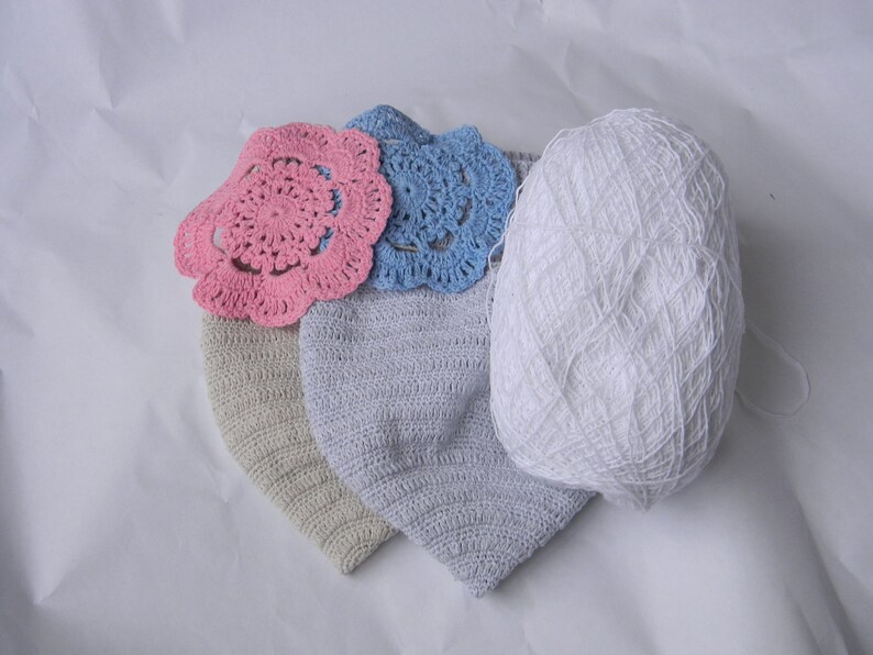 May include: Two crocheted hats, one beige and one light blue, with a pink and a blue crocheted flower on top. A ball of white yarn is also in the image.