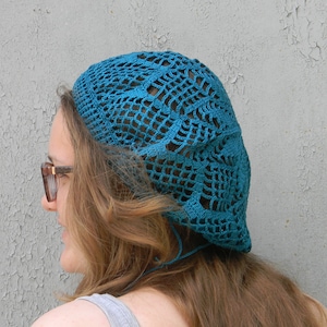 May include: A teal blue crocheted beret with a lacy design. The beret is worn on the head of a person with long brown hair.