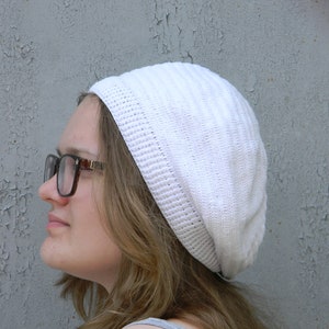May include: A woman wearing a white crocheted beanie and brown-framed glasses. The beanie has a slouchy fit and is made of a soft, textured yarn.