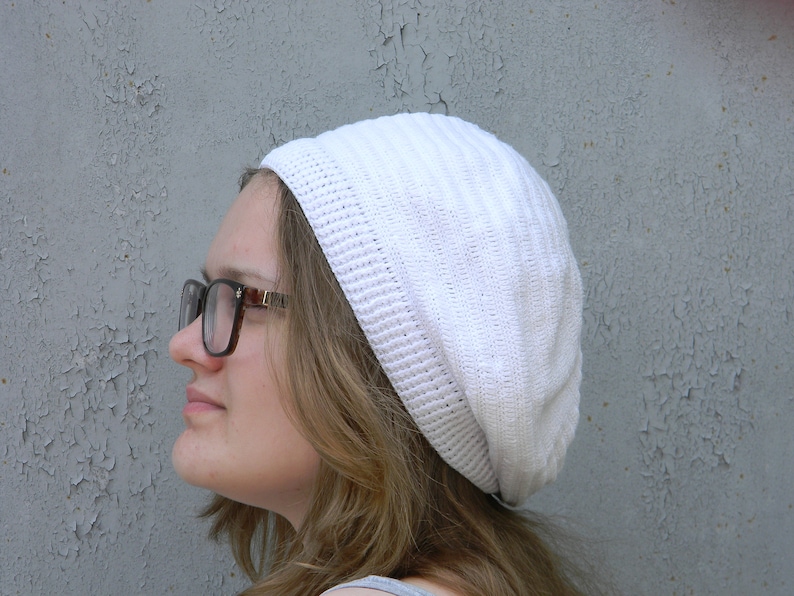 May include: A woman wearing a white crocheted beanie and brown-framed glasses. The beanie has a slouchy fit and is made of a soft, textured yarn.