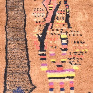May include: A brown rug with a colorful geometric design. The design features a large, abstract figure with black, red, yellow, blue, and purple stripes. The rug has a fuzzy texture.