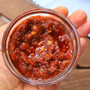 May include: A small, clear glass bowl filled with a vibrant red chili paste. The paste has a thick, textured consistency with visible pieces of chili peppers and seeds. The bowl is held in a person's hand, with a wooden surface in the background.